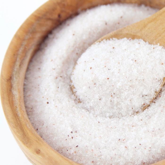 A close-up of Real Utah Salt Fine by Whole Spice Inc.—coarse, light pink salt with dark specks and trace minerals—in a wooden bowl, with a wooden spoon scooping some.