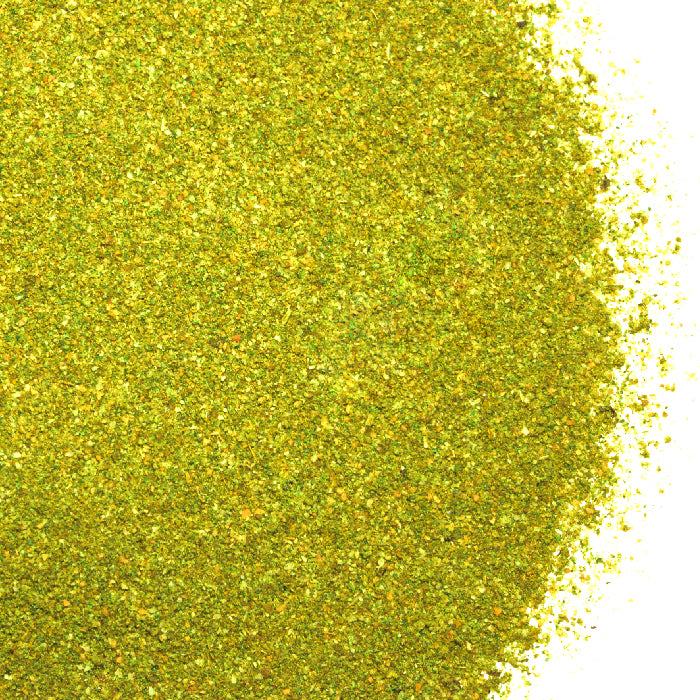 A close-up of Whole Spice Inc. Vadouvan Powder, a greenish-yellow curry blend, is spread unevenly on a white background, showcasing its fine, granular texture and colorful flecks that hint at its unique Indian spices mix.