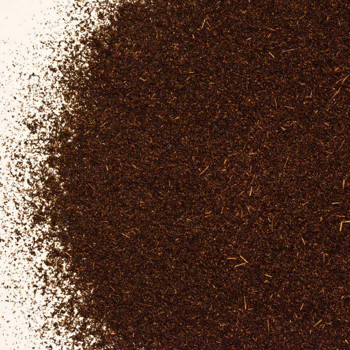 A close-up of Whole Spice Inc. Vanilla Bean Powder and loose black tea leaves on a white surface, with tea densely packed to the right and scattered alongside hints of Vanilla Bean Powder on the left.