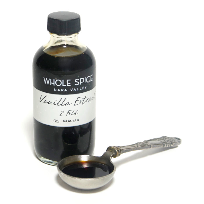 A bottle of Whole Spice Inc. Vanilla Extract 2 Fold stands upright beside a vintage silver measuring spoon filled with dark vanilla extract made from premium beans.