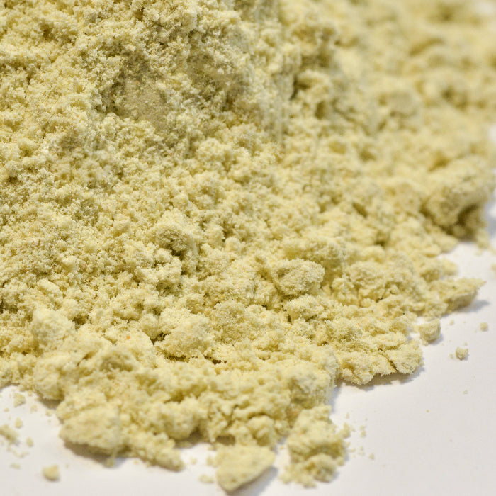 A close-up of Whole Spice Inc.'s Wasabi Blend Natural powder, a Japanese condiment, loosely scattered on a white surface. The fine pale yellow texture shows some small clumps throughout.