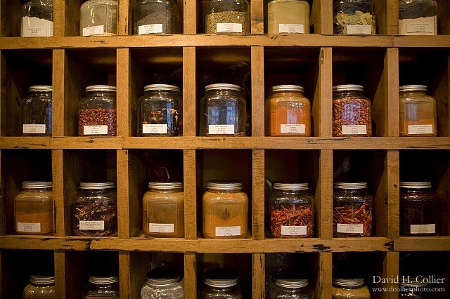 A wooden shelf holds glass jars filled with various dried herbs and spices, each labeled and neatly arranged in individual cubbyholes.