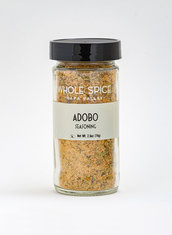 A clear glass jar with a black lid labeled “Adobo Seasoning” by Whole Spice Inc. contains a blend of ground spices ideal for meat rubs, with NO MSG featured on the label. Net weight: 2.6 oz (74g). The background is plain white.
