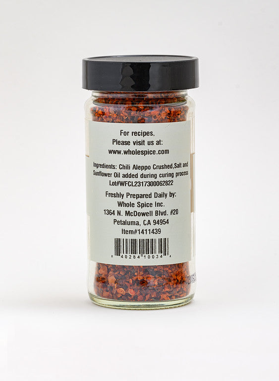 A clear jar with a black lid contains vibrant Aleppo Chili Crushed by Whole Spice Inc. The white label details ingredients, preparation, and contact info—ideal for fans of Mediterranean cuisine.