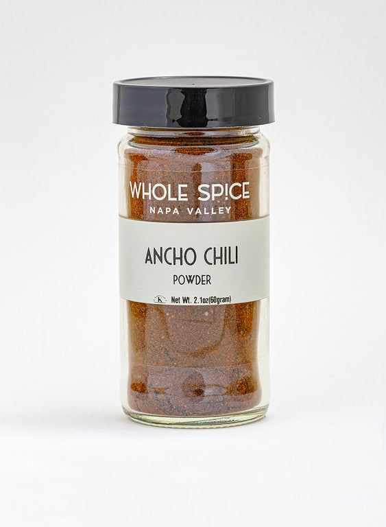 A glass jar with a black lid holds Whole Spice Inc. Ancho Chili Powder, Net Wt. 2.1oz (60g), ideal for Mexican dishes and mole. The jar is photographed against a plain white background.