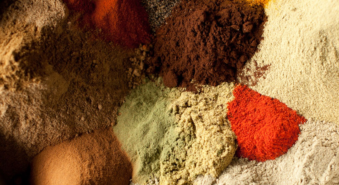 A close-up of various colorful ground spices arranged in small piles, including shades of brown, red, yellow, green, and orange, creating a vibrant and textured pattern.