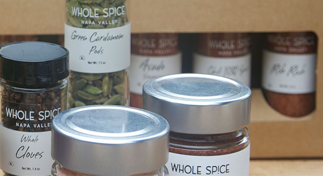 Assorted Whole Spice Napa Valley jars, including green cardamom pods and whole cloves, are displayed in the foreground with more spice jars in a cardboard box in the background.