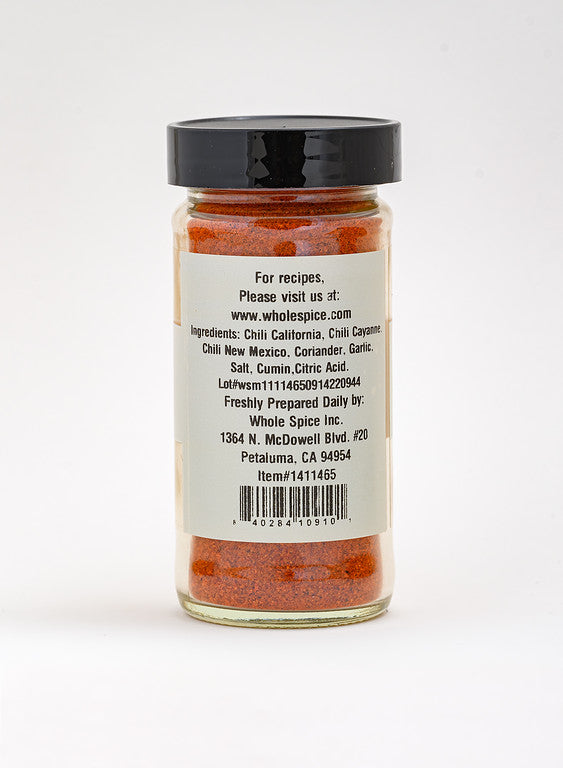 A clear jar of Whole Spice Inc. Harissa Spice with a black lid holds orange-red seasoning. The back label features ingredients, website, address, and details in bold black text.