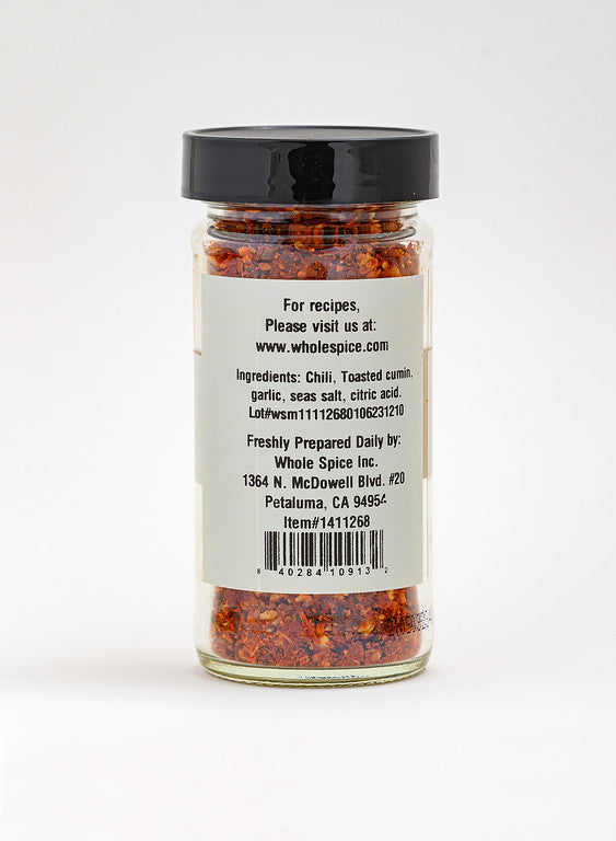 A clear glass jar from Whole Spice Inc. labeled "Harissa Spice Coarse" features a black lid and back label with ingredients, website, and address. Inside is a red, coarse Moroccan Harissa chili blend with visible seeds and flakes.