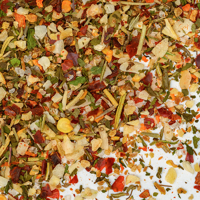 A close-up of Whole Spice Inc.'s Lamb Rub shows a colorful blend of dried herbs and spices, with vibrant red, green, and yellow pieces—like onions, peppers, and Mediterranean seasonings—scattered on a white surface.