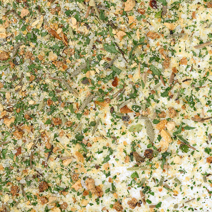 A close-up of Whole Spice Inc. Lemon Herb Seasoning shows chopped green herbs, dried onion, garlic flakes, and spices scattered on a white background.