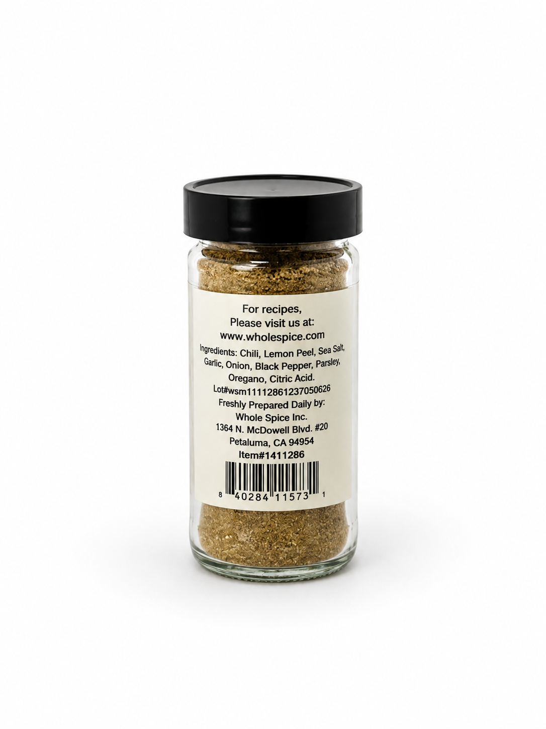 A glass spice jar from Whole Spice Inc., topped with a black lid, displays a label for the Shawarma Spice Mix featuring ingredients such as chili, lemon peel, and sea salt. The label also provides a website, contact address, barcode, and preparation details for this aromatic blend inspired by traditional Middle Eastern spices.
