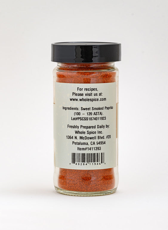 A clear spice jar with a black lid contains red Sweet Smoked Paprika from Whole Spice Inc. The label lists ingredients, lot number, company address, website, and item number—featuring authentic Spanish paprika.