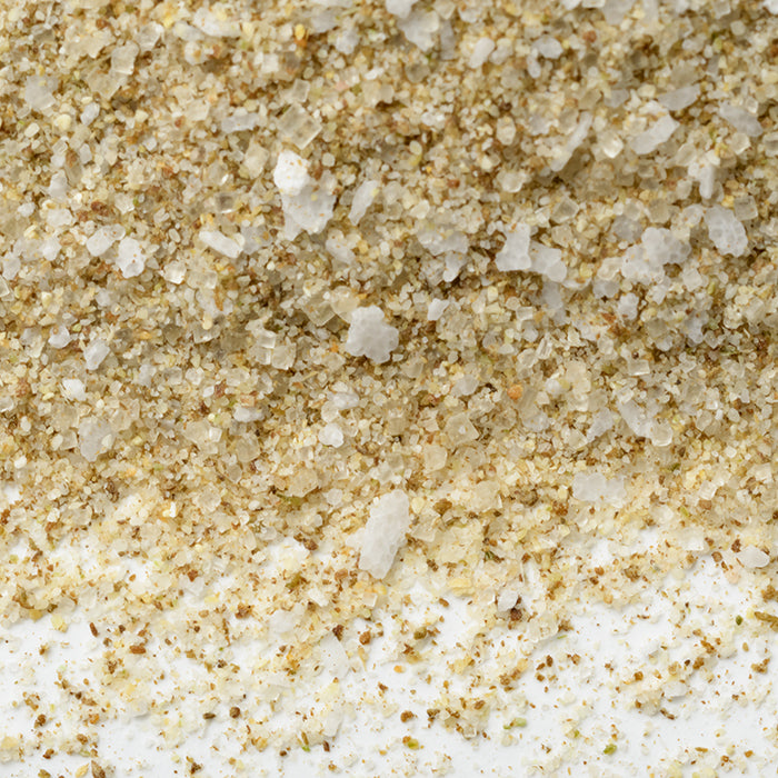 Close-up of Le Beau Monde by Whole Spice Inc., a savory spice blend with coarse, light brown and white grains and visible herb flecks, spread unevenly across a white surface.