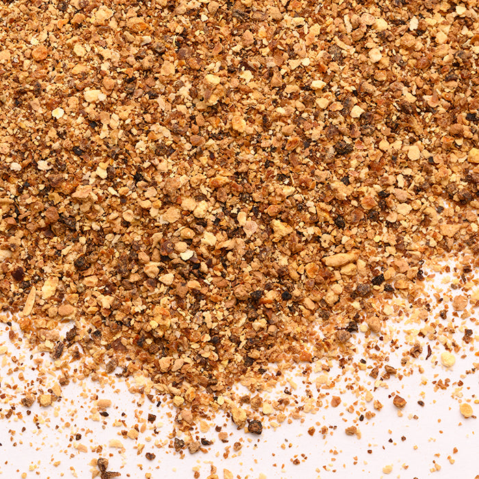 A close-up of scattered Orange Peel Granules by Whole Spice Inc. on a white background, displaying their coarse, golden-brown texture and hints of citrus seasoning.