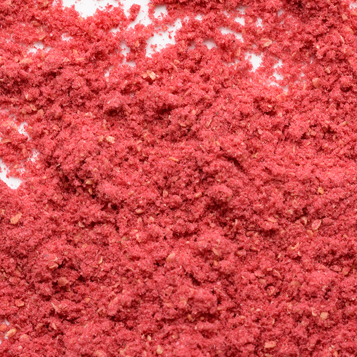 A close-up of Whole Spice Inc. Raspberry Fruit Powder, showing its pinkish-red, coarse texture. The slightly uneven powder is spread out, filling the entire frame.