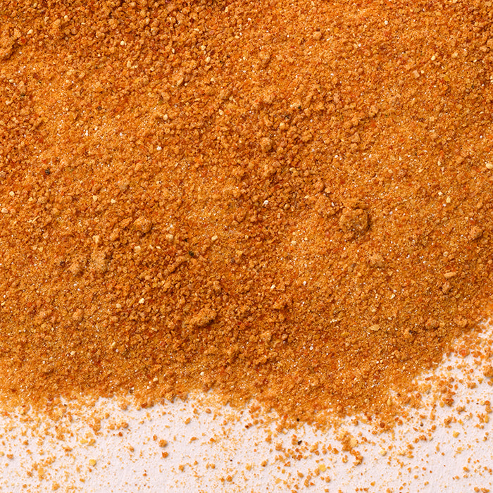 A close-up of Jalapeño Red Chili Powder by Whole Spice Inc. shows orange-brown granules scattered on a white surface, highlighting its textured, powdery appearance with small, uneven particles.