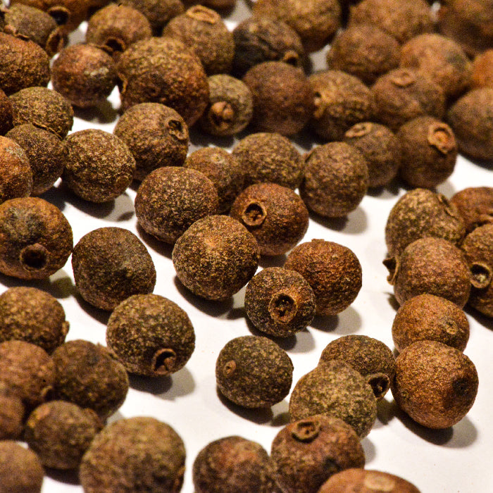 A close-up of Whole Spice Inc. Allspice Whole shows scattered, round brown berries with a rough texture and slight size variations on a white surface. Also known as Jamaica pepper, these dried berries are aromatic and flavorful.
