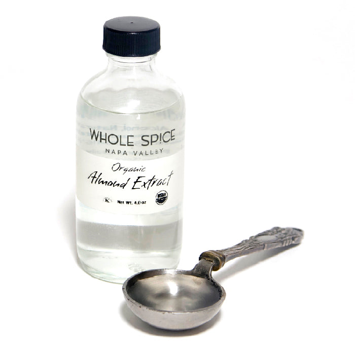 A clear glass bottle of Almond Extract by Whole Spice Inc. sits beside an ornate metal measuring spoon on a white surface, ready to add rich almond flavor to your favorite desserts.