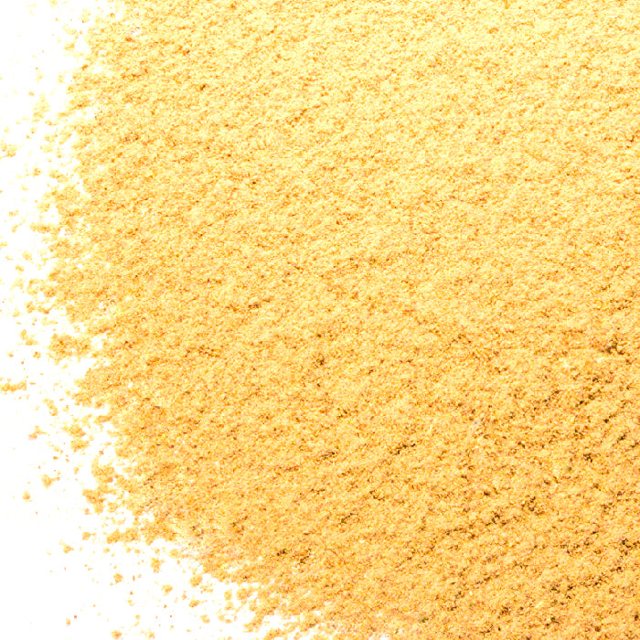 A pile of fine, yellowish Amchur Powder by Whole Spice Inc.—a tangy Indian spice made from dried mangoes—lies on a white surface, densely concentrated on the right and gently scattering leftward.
