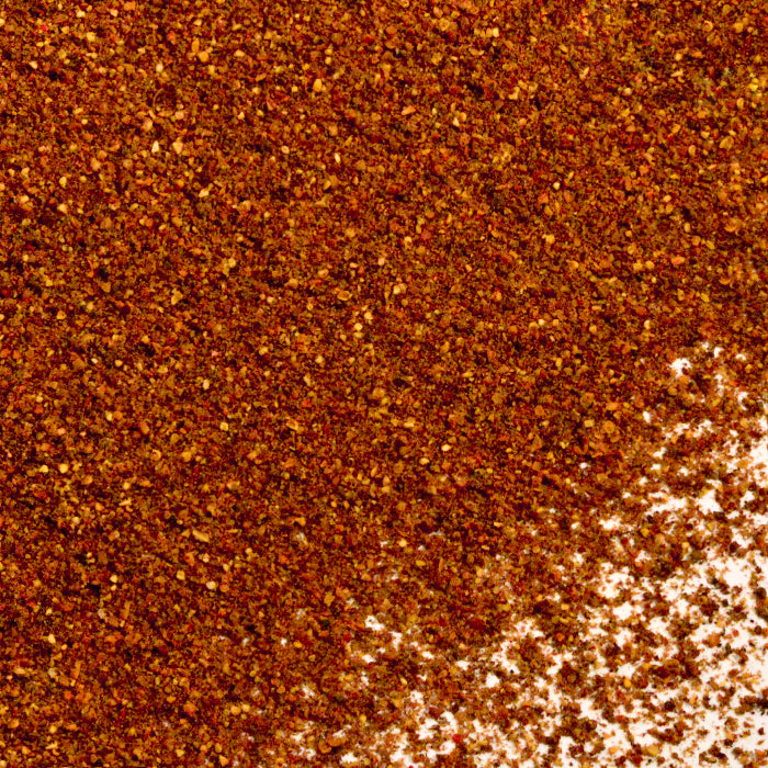 A close-up of Whole Spice Inc. Anardana Powder—reddish-brown and finely ground—scattered on a white surface, with some areas more densely packed, highlighting the essence of dried pomegranate seeds.