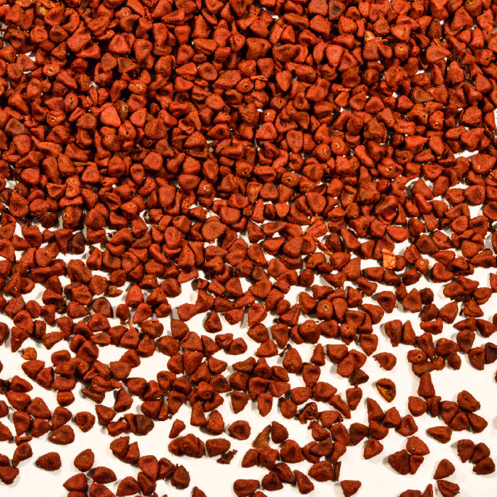 A dense layer of reddish-brown Annatto Seeds by Whole Spice Inc. is scattered against a white background, with some seeds dispersed more sparsely toward the bottom, highlighting their value as a natural coloring.