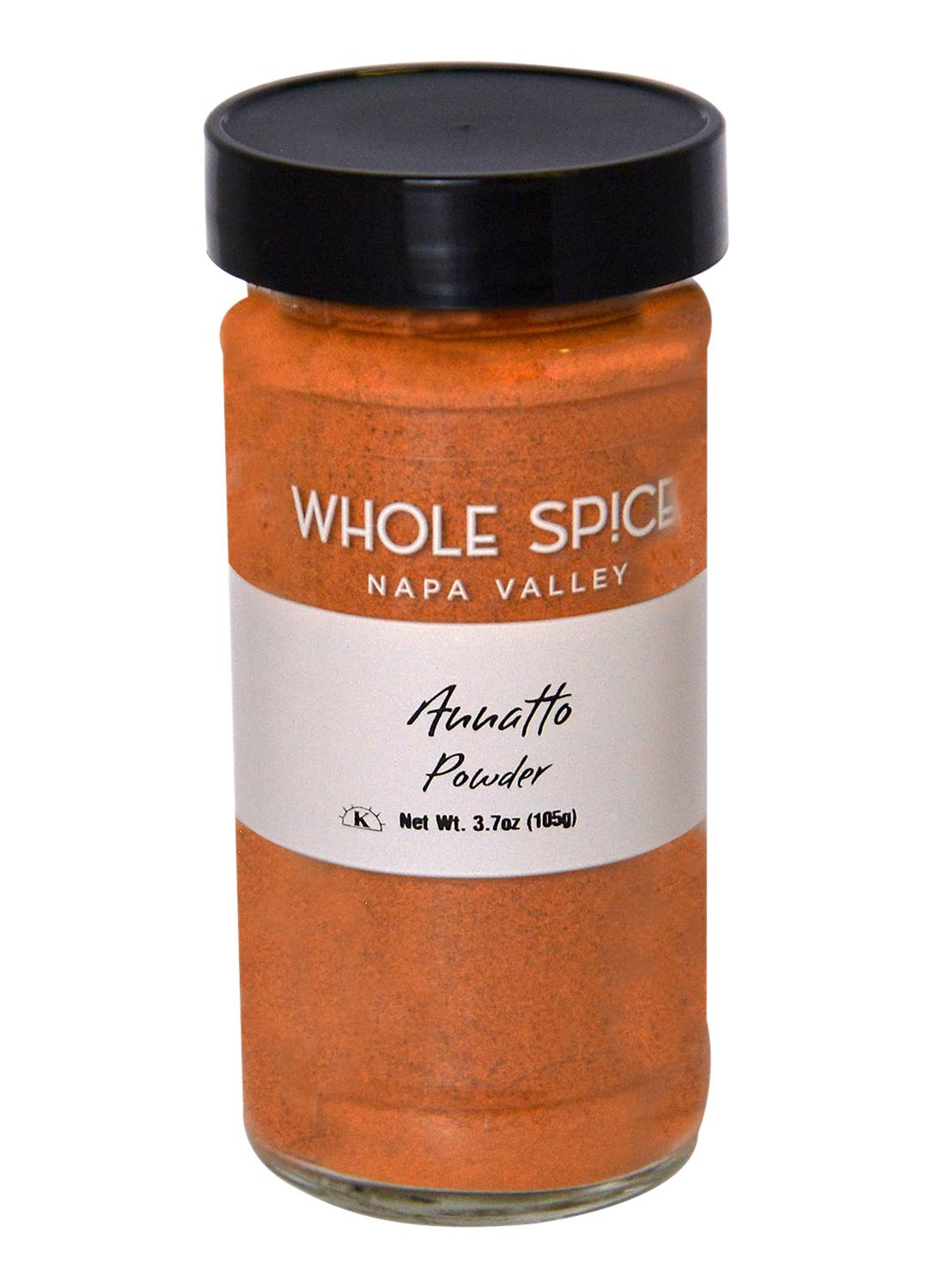 A glass jar of Whole Spice Inc. Annatto Powder, filled with vibrant orange-red spice from natural annatto seeds, features a black lid and a white label showing the product name and weight.