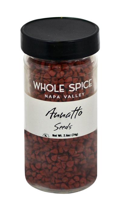 A clear glass jar with a black lid labeled Whole Spice Inc. Annatto Seeds contains small reddish seeds used as natural coloring. Net weight: 2.6 oz (74g).