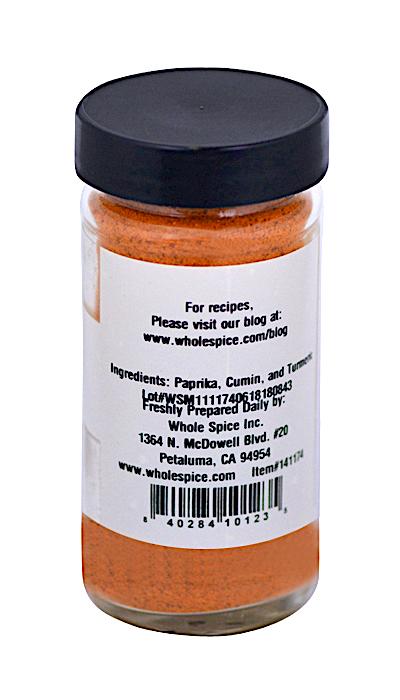A clear spice jar from Whole Spice Inc. labeled "Basic Curry Powder" with a black lid, contains an orange blend of paprika, cumin, and turmeric. The label shows ingredients, contact info, and barcode; front label not visible.