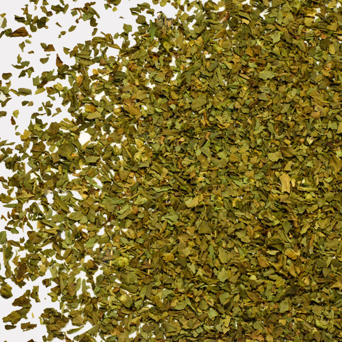 A close-up of dried and crushed Whole Spice Inc. Basil Leaves scattered on a white surface, showcasing their vibrant green color and freshness.