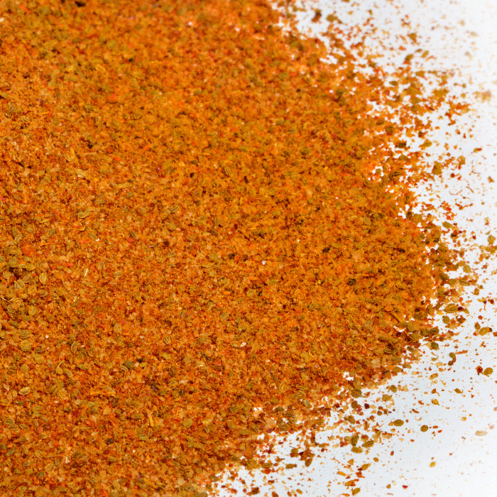 A close-up of Bay Seafood Seasoning by Whole Spice Inc., finely ground and scattered on a white surface, shows orange and brown colors with visible flecks of herbs and seasonings.