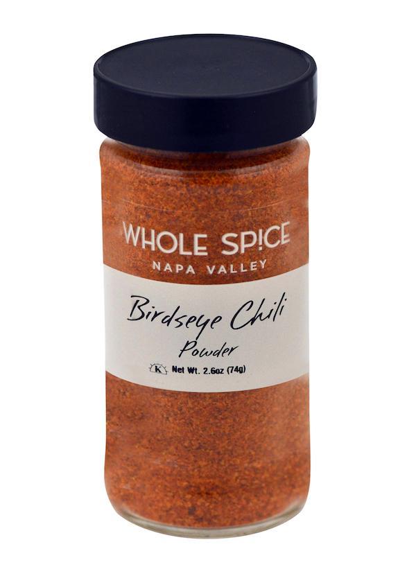A clear glass jar with a black lid labeled Whole Spice Inc. Bird's Eye Chili Powder contains fiery reddish-orange chili powder. Net weight: 2.6 oz (74g).