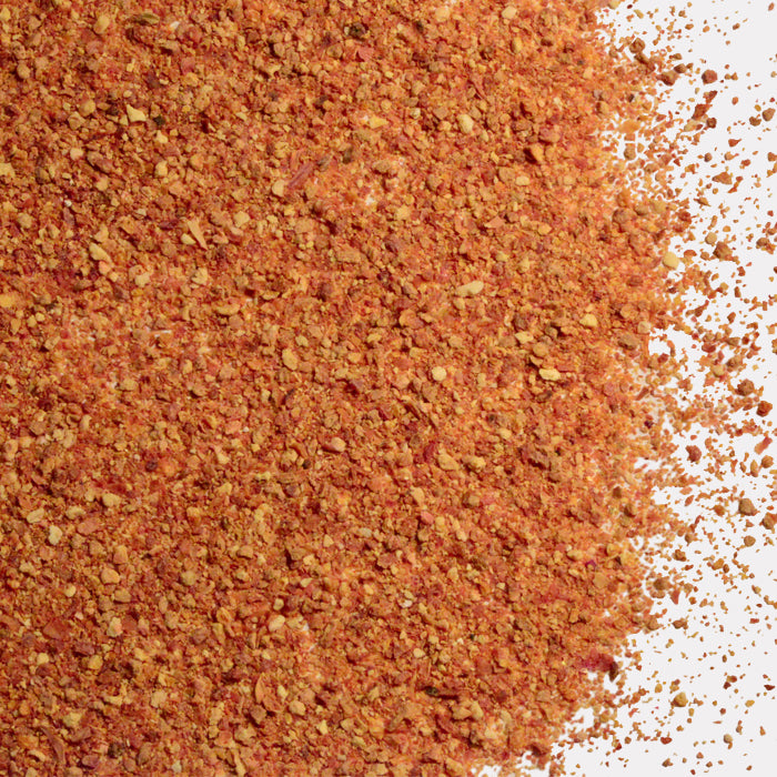 A close-up of Whole Spice Inc. Blood Orange Peel Granules, a coarse, reddish-brown seasoning with visible pieces, scattered on a white surface.