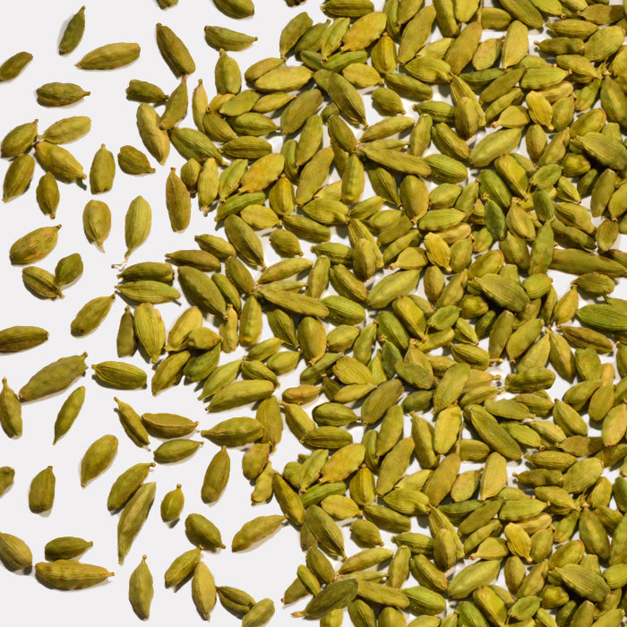 Whole Spice Inc. Green Cardamom Pods, a key ingredient in Indian spiced tea, are scattered on a white background—some grouped together and others spread apart.