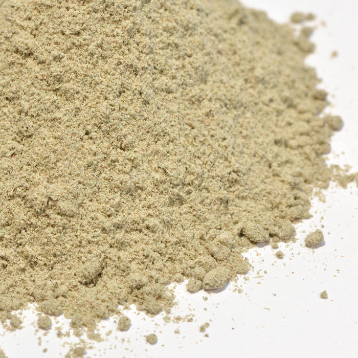 A close-up of a small mound of Whole Spice Inc. Green Cardamom Powder Organic, finely ground and light beige, sits on a white surface with some clumps and loose grains scattered around the pile.