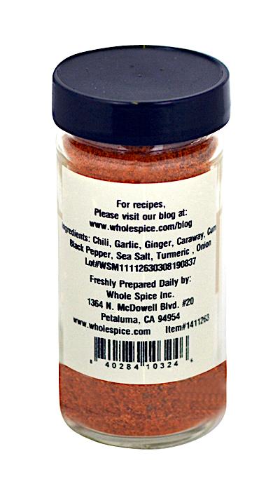 A clear spice jar with a black lid contains Whole Spice Inc.'s Charmoula Seasoning—a red-orange blend featuring chili, garlic, and ginger—ideal for Moroccan dishes. The label includes contact details and blog info for Whole Spice Inc.