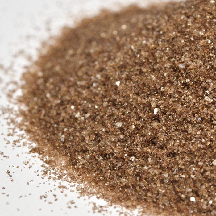 A close-up of Whole Spice Inc. Cherrywood Smoked Sea Salt, featuring coarse, brown, glittery granules scattered on a white surface and evoking rich wood-fired flavor.