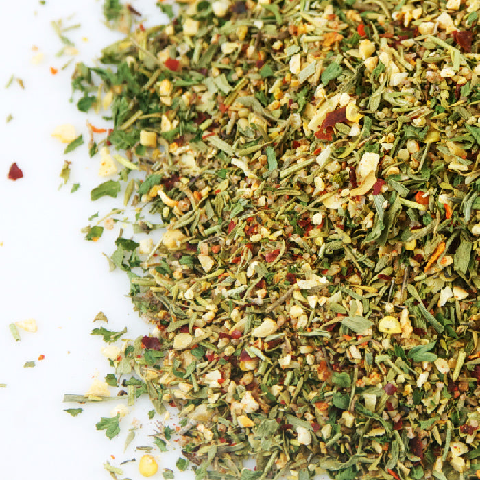 A close-up of Whole Spice Inc. Rotisserie Chicken Rub showcases its colorful, coarse blend of dried herbs and spices with green, red, and yellow flecks scattered on a white surface.