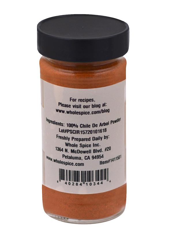 A clear jar with a black lid from Whole Spice Inc. contains Chili De Arbol Powder, a reddish-orange Mexican chili powder. The back label states "100% Chili De Arbol powder" and includes company info plus a barcode at the bottom.