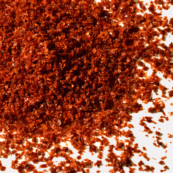 Whole Spice Inc. Marash Chili Crushed is a vibrant red-orange chili flake, scattered across a white surface with loose flakes around the main pile—a flavorful staple in Middle Eastern cuisine.