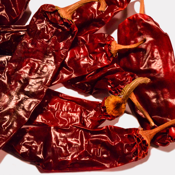 A close-up of Whole Spice Inc. California Chili Whole shows several dried red chilies with wrinkled skins and stems, overlapping on a light surface—ideal for adding authentic flavor to Mexican dishes.