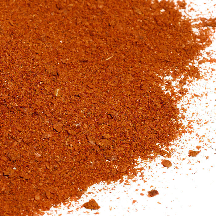 A close-up of Chili Spice Blend - Fiery Hot by Whole Spice Inc. shows a reddish-brown, fine powder with small chunks on a white surface, highlighting its bold heat and vibrant texture.