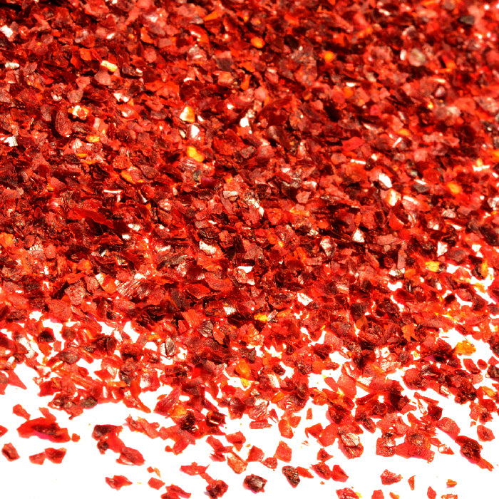 Close-up of vibrant California Chili Crushed flakes by Whole Spice Inc. scattered on a white background, showcasing their shiny, textured, and irregular shapes.