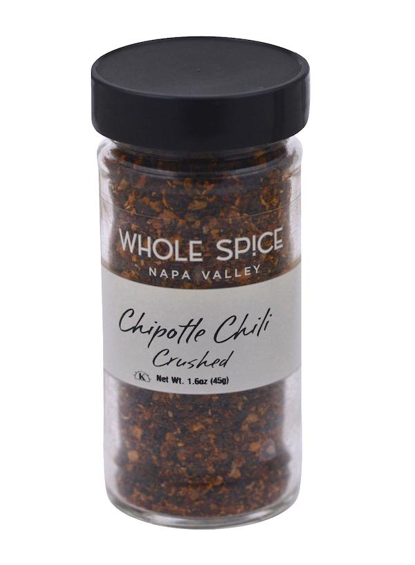 A clear glass jar with a black lid holds crushed chipotle chili flakes made from smoked jalapeños. The label says Whole Spice Inc., Chipotle Chili Crushed, Net Wt. 1.6oz (45g).