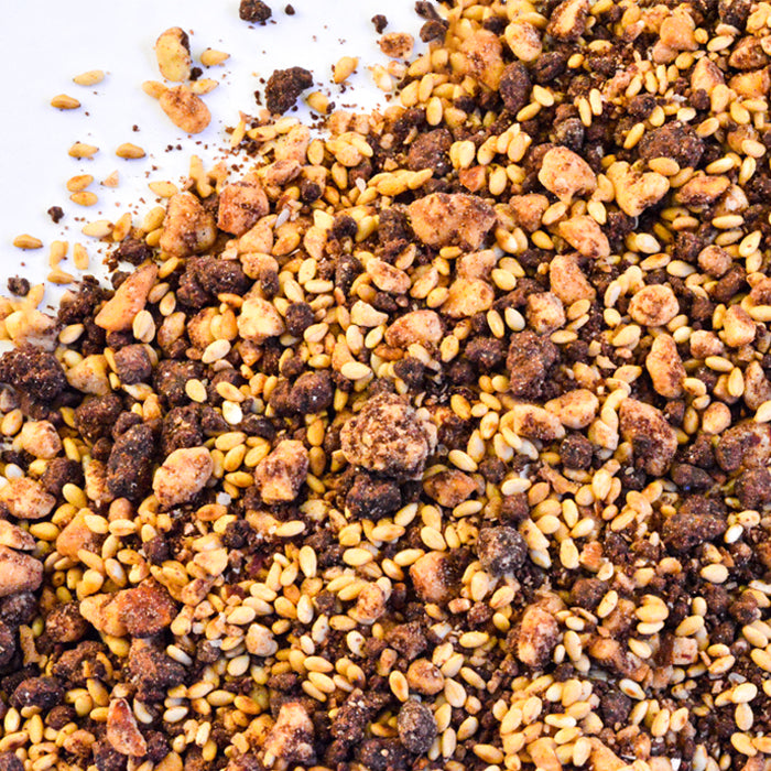 A close-up of Whole Spice Inc. Chocolate Dukka, featuring small golden sesame seeds, toasted nuts, and brown crumbly clusters scattered on a white surface.