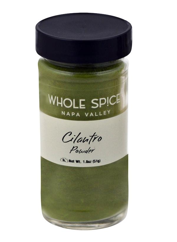 A glass jar with a black lid labeled Whole Spice Inc. Cilantro Powder holds 1.8 oz (51g) of green powdered cilantro leaves, a staple herb in Mexican cuisine.