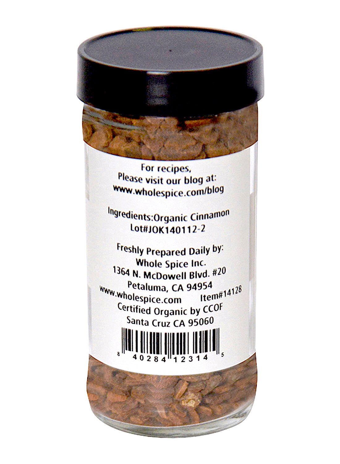A clear glass jar with a black lid holds "Cinnamon Chips Organic" by Whole Spice Inc. The label features the ingredient, company info, certification, and a barcode; contents are organic cinnamon bark chips used in traditional Chinese medicine.