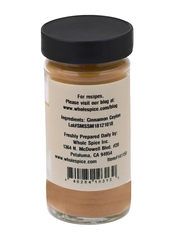 A glass jar with a black lid contains Cinnamon Powder Ceylon by Whole Spice Inc., made from authentic Sri Lanka cinnamon. The label lists ingredients, company address, website, and barcode. This prized spice is perfect for baking.