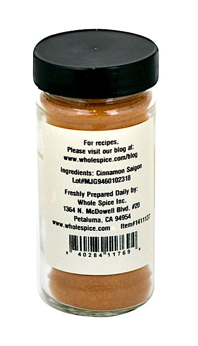 A glass spice jar of Whole Spice Inc. Cinnamon Powder Saigon with a black lid is shown from the back; its label lists Saigon cinnamon as the ingredient, company contact details, address, website, and a barcode at the bottom.