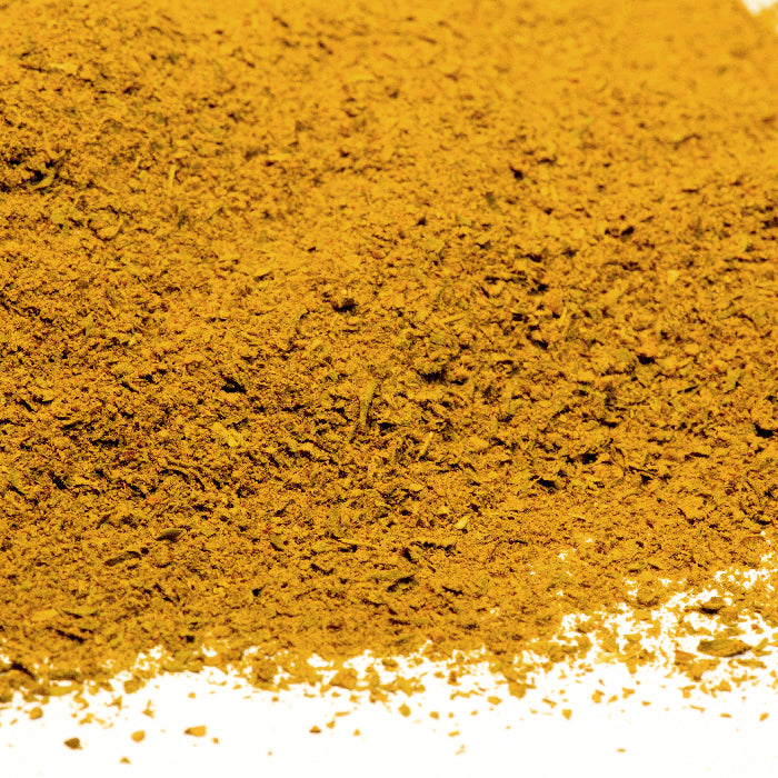 A close-up of Whole Spice Inc.'s Couscous Spice Mix, a ground yellowish-brown powder, scattered on a white surface and ideal for Moroccan couscous or curry dishes.